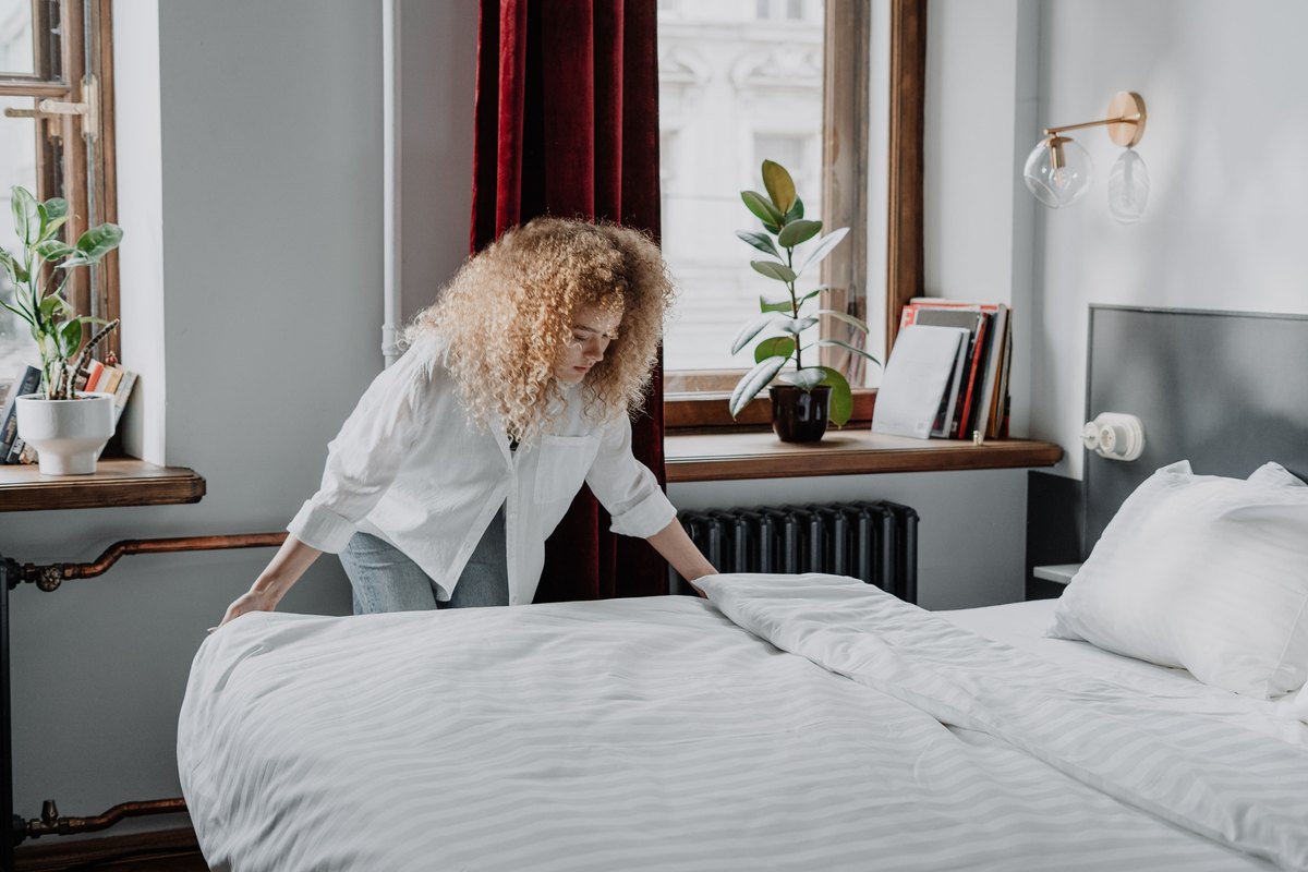 Woman with curly hair in a white shirt leaning over a bed to smooth a white striped duvet